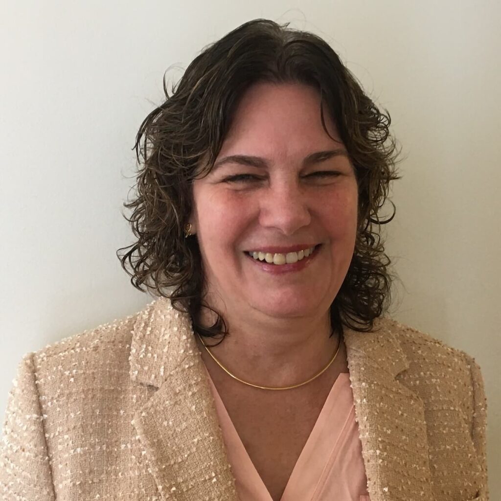 Headshot of DeAnn Elliott, a woman with wavy brown hair, smiling and wearing a light pink blouse under a beige textured blazer.